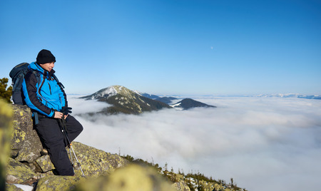Tourist hiker with backpack and trekking poles resting on steep rocky mountain slope on background of blue sky, foggy valley filled with white clouds and snowy mountain tops.の写真素材