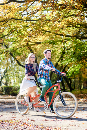 Young tourist pair, handsome bearded man and attractive blond long-haired woman cycling together tandem double bike by sunny alley with golden leaves on tall trees background in the fallの写真素材