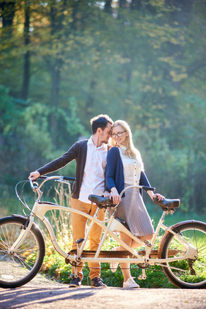 Young happy couple tourists, bearded man and attractive woman close together at tandem double bicycle outdoors in summer park or forest on blurred sunny green trees dense foliage background.の写真素材
