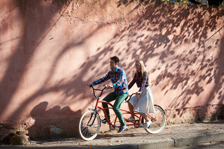 Young happy tourist couple in casual clothing, bearded man and blond woman in glasses cycling tandem bike along empty paved sunny sidewalk on background of high red crackled plastered wall.の写真素材