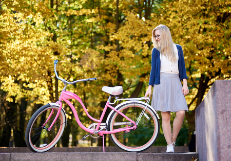 Young active blond long-haired woman in skirt and blouse standing with pink lady bicycle on lit by autumn sun bright blurred colorful golden bokeh park trees foliage background.の写真素材