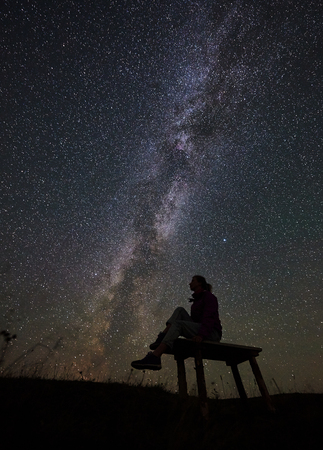 Dark silhouette of female hiker having a rest at summer night in the mountains. Happy woman traveller sitting on a bench, enjoying view of night sky full of stars and Milky way.の写真素材