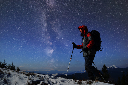 Lonely experienced male tourist with trekking sticks during winter hiking getting mountain peak in darkness. Silhouette in mountains and amazing view. Blue starry sky and Milky Way on background.の写真素材