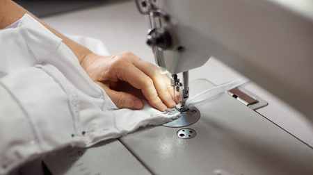 Female hands stitching white fabric on professional manufacturing machine at workplace. Seamstress hands holding lace textile for dress production. Blurred background. Close up view of sewing process.の写真素材