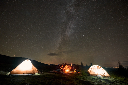 Three tourist tents on mountain valley and company of happy people, men and women with guitar by burning bonfire on copy space background of dark starry sky with bright Milky Way constellation.の写真素材