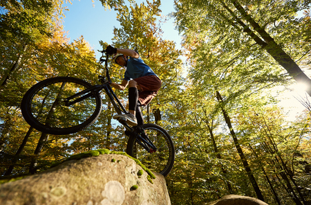 Professional sportsman cyclist riding on back wheel on trial bicycle, man biker making acrobatic stunt on the edge of big boulder in the forest on sunny day. Concept of extreme sport active lifestyleの写真素材