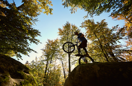 Professional sportsman biker balancing on back wheel on trial bike, cycler making acrobatic trick on the edge of big boulder in the forest on sunny day. Concept of extreme sport active lifestyleの写真素材