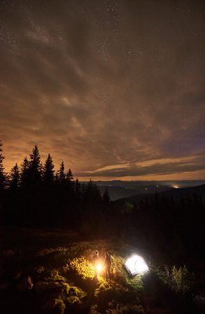 Vertical shot of night summer camping in the mountains. Top view of happy couple hikers having a rest together, standing beside campfire and glowing tourist tent. On background cloudy sky with stars.の写真素材