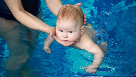 Active baby swimming in womans hands in swimming pool. Cropped female hands holding carefully babys...