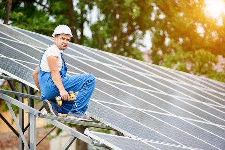 Young smiling electrician worker sitting on almost finished stand-alone solar photo voltaic panel system on bright sunny green tree background. Alternative energy concept.の写真素材