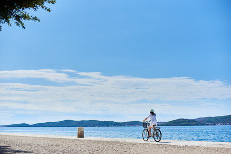 Back view of woman in white summer closing and hat riding a bicycle along stony sidewalk under clear blue sky. Sparkling sea water and mountains view on opposite shore on background.の写真素材