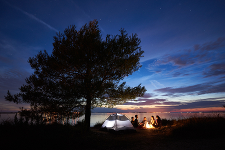 Group of five young tourists sitting on sea shore around campfire near tent under big tree and blue evening sky with first stars at sunset. Tourism, friendship camping and beauty of nature concept.の写真素材