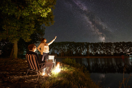 Young couple travellers resting near campfire on a lake shore. Man sitting on chair, woman pointing to evening sky full of stars and Milky way above still water and forest on background. Night campingの写真素材