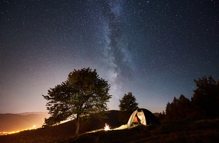 Tourist camping near forest at night. Illuminated tent and bonfire under magical night sky full of stars and Milky way. On background incredibly beautiful starry sky, mountains and luminous townの写真素材