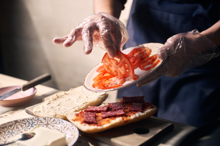 Chef mastery hands holding white plate with slices of cheese and sausage. Putting pieces of tomato on bread greased with red sauce for cooking panini. Wooden working surface. Blurred background.の写真素材