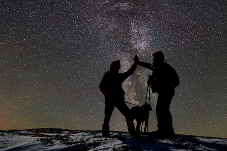 Adult experienced skiers with dog rejoicing after reaching goal on snow-covered mountain peak during night ski-tour. Silhouettes in mountains. Starry sky before sunrise and Milky Way on background.の写真素材