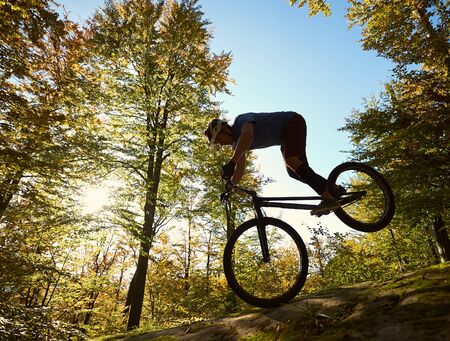 Professional cyclist balancing on front wheel on trial bike. Courageous sportsman rider making acrobatic trick on the edge of big boulder in the forest on sunny day. Concept of extreme sportの写真素材