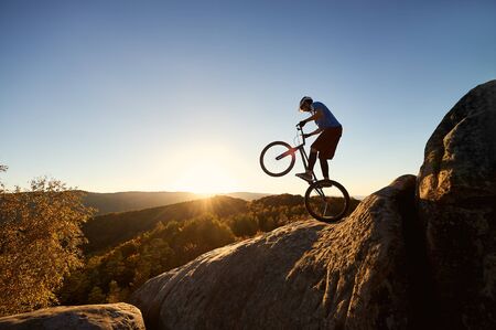 Fearless cyclist standing on back wheel on trial bicycle. Professional sportsman rider balancing on the edge of big boulder on the top of mountain at sunset. Concept of extreme sportの写真素材
