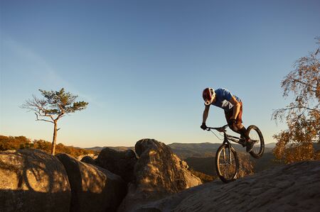 Professional cyclist balancing on front wheel on trial bike. Sportsman rider making acrobatic trick on the edge of big boulder on the top of mountain. Concept of extreme sport active lifestyleの写真素材