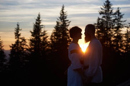 Silhouettes of happy couple standing on the top of mountain in the evening. Bearded man and pretty woman looking to each other. Beautiful sunset, cloudy sky and forest on blurred background.の写真素材