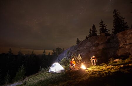 Night camping in the mountains. Young couple hikers having a rest together, sitting beside campfire and illuminated tourist tent. On background big boulder, forest and beautiful night starry sky.の写真素材