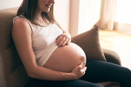 Cropped view of pregnant woman in white shirt sitting on sofa at home, embracing belly and smiling. Happy mother enjoying pregnancy and expecting little baby. Concept of happiness.の写真素材