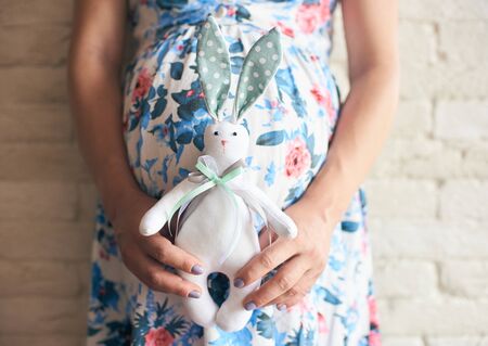 Cropped view of pregnant woman in flowery dress holding cute toy bunny. Future mother in anticipation of baby. Concept of pregnancy and expectation.の写真素材