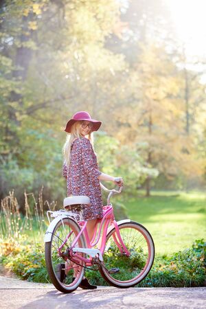 Slim blond fashionable long-haired attractive girl in glasses, short dress and pink hat at lady bicycle outdoors on paved sunny summer park alley on beautiful foggy green and golden trees background.の写真素材