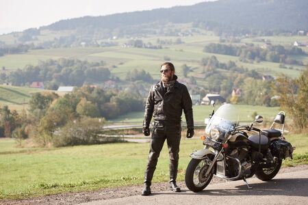 Bearded cool tall athletic sportive motorcyclist in dark sunglasses and black leather clothing, standing at modern cruiser motorbike on background of green rural landscape and woody hills.の写真素材
