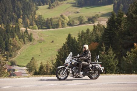 Side view of bearded motorcyclist in helmet, sunglasses and black leather clothing riding cruiser motorbike along narrow asphalt path on sunny summer day on background of tall trees and greeen grass.の写真素材