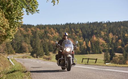 Bearded motorcyclist in helmet, sunglasses and black leather clothing riding cruiser motorcycle along empty asphalt road on bright sunny autumn day.の写真素材