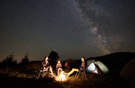 Happy family hikers resting together in the mountains, sitting on chairs beside camp, bonfire and...