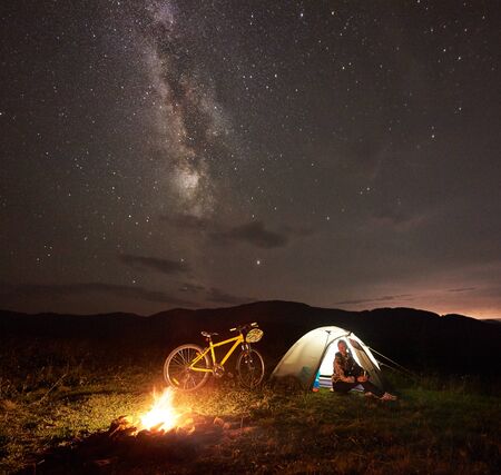 Young woman tourist having a rest at night camping near burning campfire, illuminated tourist tent, mountain bicycle under amazing beautiful evening sky full of stars and Milky way. Astrophotography