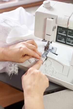 Female hands edging white lace fabric during sewing process on professional overlock machine at workplace. Seamstress hands holding lace textile for dress production. Blurred background. Close up viewの写真素材