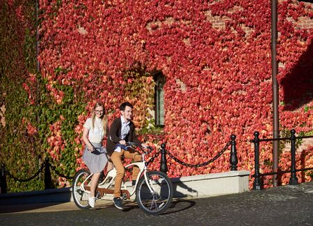 Young happy tourist couple, handsome man and pretty long-haired blond woman in glasses cycling tandem bike on bright sunny autumn day on background of brick wall fully overgrown with red ivy leaves.の写真素材
