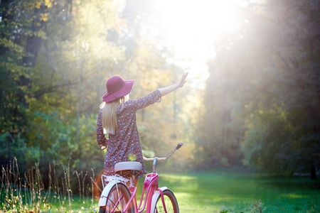 Back view of slim blond fashionable long-haired attractive girl in short dress and pink hat with raised arm at lady bicycle on paved summer park alley, enjoying beautiful evening sunsetの写真素材