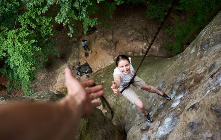 Happy woman climbing rock. Carefree hiker smiling her friend, hanging on rope and stretching out hand, asking for help. Man helping his friend to climb rock. Helping hand. Teamwork concept. Top view.の写真素材