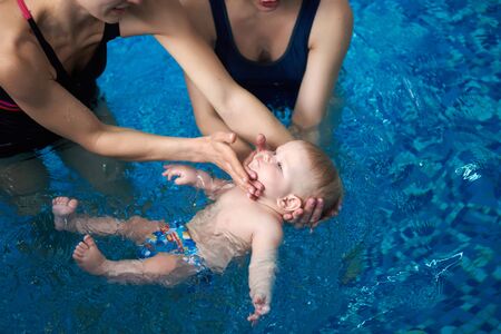 Cropped view of two women standing in pool, trying carefully keeping baby on water. Cute calm little child in swimming nappies lying on his back in blue water. Kids swimming healthy lifestyle conceptの写真素材