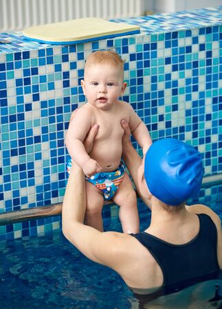 Portrait of cute infant boy looking in camera, sitting and resting with mother in paddling swimming pool. Mom preparing future swimmer sportsman. Parents spending active time with their babyの写真素材