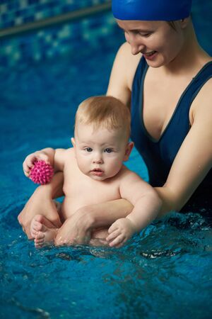 Vertical portrait view of baby holding toy balls and sitting in his mothers hands in clear blue water of swimming pool during wellness treatments. Children water activity and fitness with parentsの写真素材