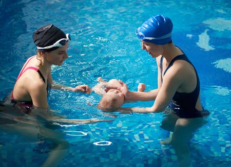 Side view of mother and female young instructor carrying out swimming lessons for parent and baby. Mom and her newborn child at infant swimming class. Baby floating lying on his back in blue waterの写真素材
