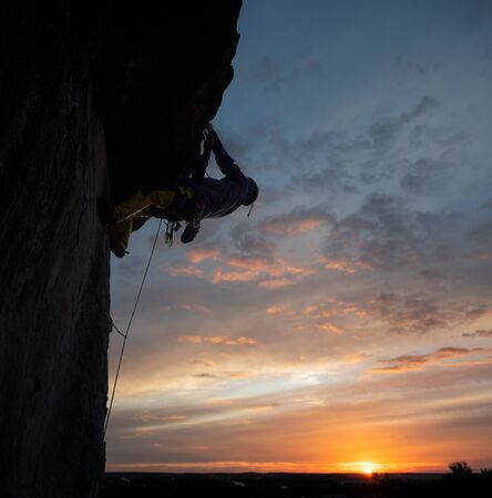 Back low angle view of male climber on rocky wall at sunset conquering the top of steep cliff. Evening sky with sunlight on horizon on background. Active outdoors extreme lifestyle concept. Copy spaceの写真素材