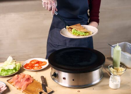 Chefs female hands keeping white plate and putting in with spatula sandwich crepe with green salad and rolled up in triangle. Dish serving process. Appetizing hot fried food for lunch. Close up view.の写真素材