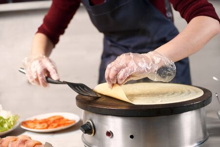 Cropped woman chef hands in gloves turn over big pancake with spatula on electric round cooktop while baking process. Lettuce, salmon and tomato slices in plate on blurred light background. Front viewの写真素材