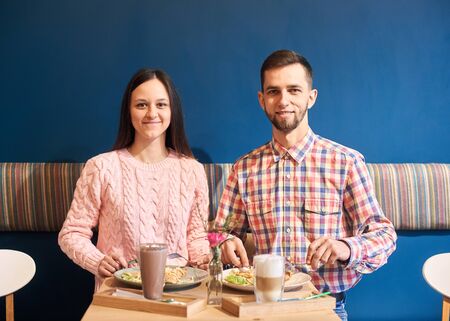 Portrait of stylish brunette happy young couple sitting one by one at small table and looking at camera. Enjoying appetizing lunch food and beverages in restaurant. Blue decorated wall on background.の写真素材