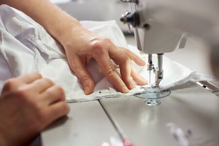 Close up side top view of cropped female hands sewing white fabric on professional manufacturing machine at seamstress workplace. Tailor hands holding textile for dress production. Blurred backgroundの写真素材