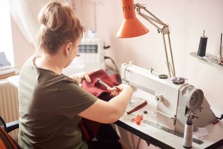 Back view of tailor laboring at working place in atelier. Female hands using scissors for cutting off cloth piece in sewing process. Sewing machine, ruler, scissors, table lamp, threads on background.の写真素材