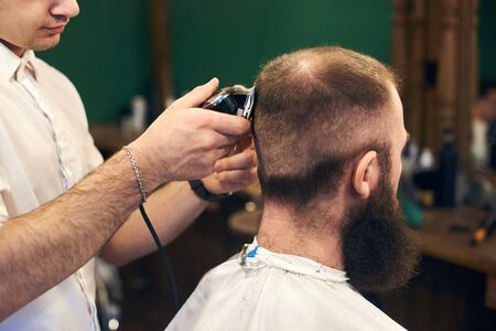 Perfect trimming. Back side close-up view of young bearded man getting haircut by masterful barber with electric razor while sitting in hairdresser chair. Contemporary service in barbershops.の写真素材