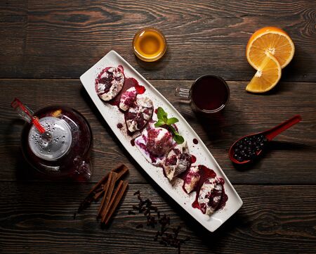 Top view of delicious Asian dessert in long white plate with glass kettle full of tea, honey, orange, glass of tea, spoon with blueberries, cinnamon sticks and carnation scattered on wooden dark tableの写真素材
