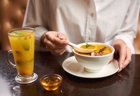 Crop of woman in white blouse holding in hands white bowl with orange cream soup. Female eating in restaurant first dish and tea with mint and honey near. Concept of healthy food.の写真素材